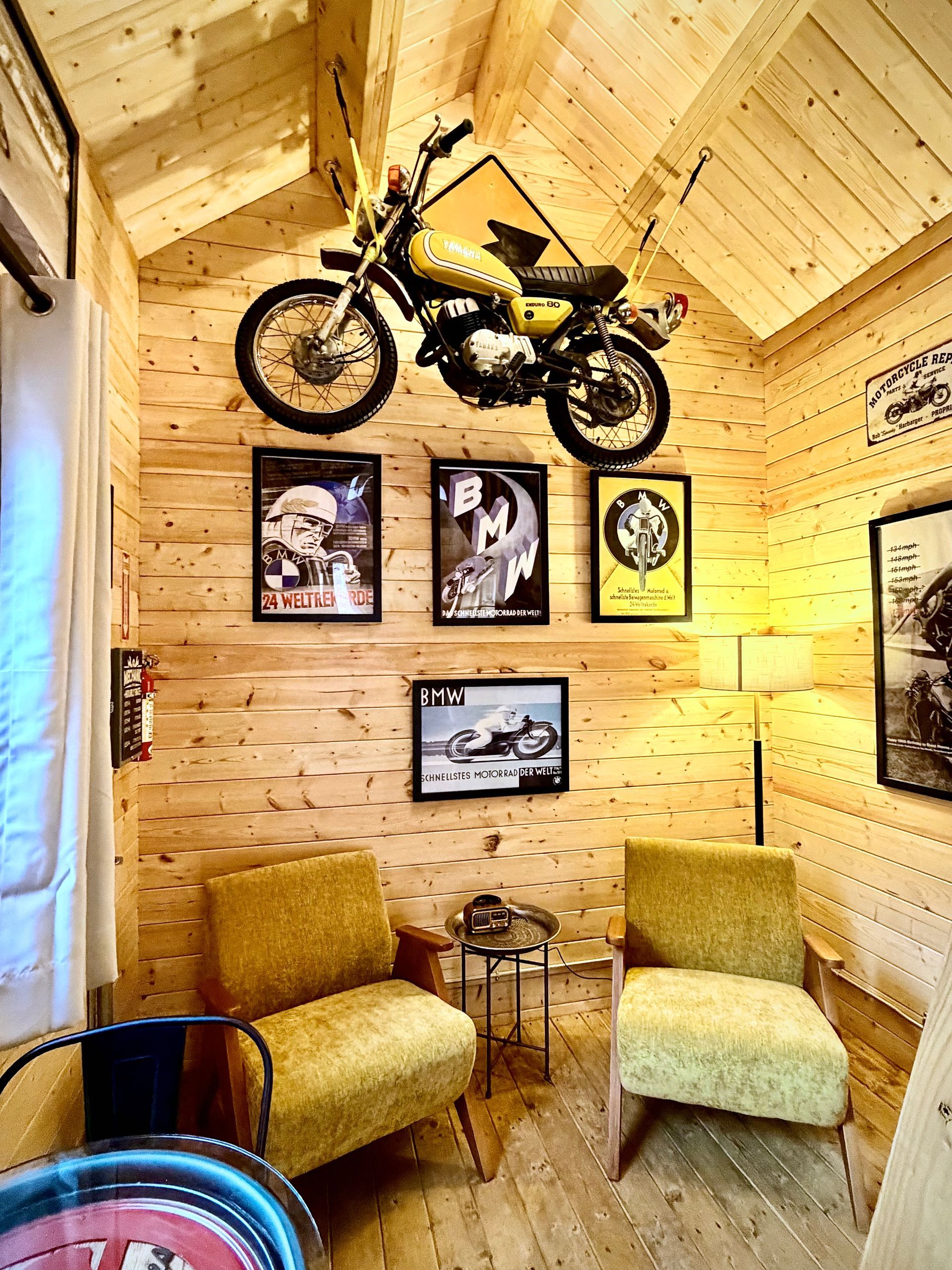 Wood-paneled room with a wall-mounted motorcycle above two chairs and framed art, warmly lit