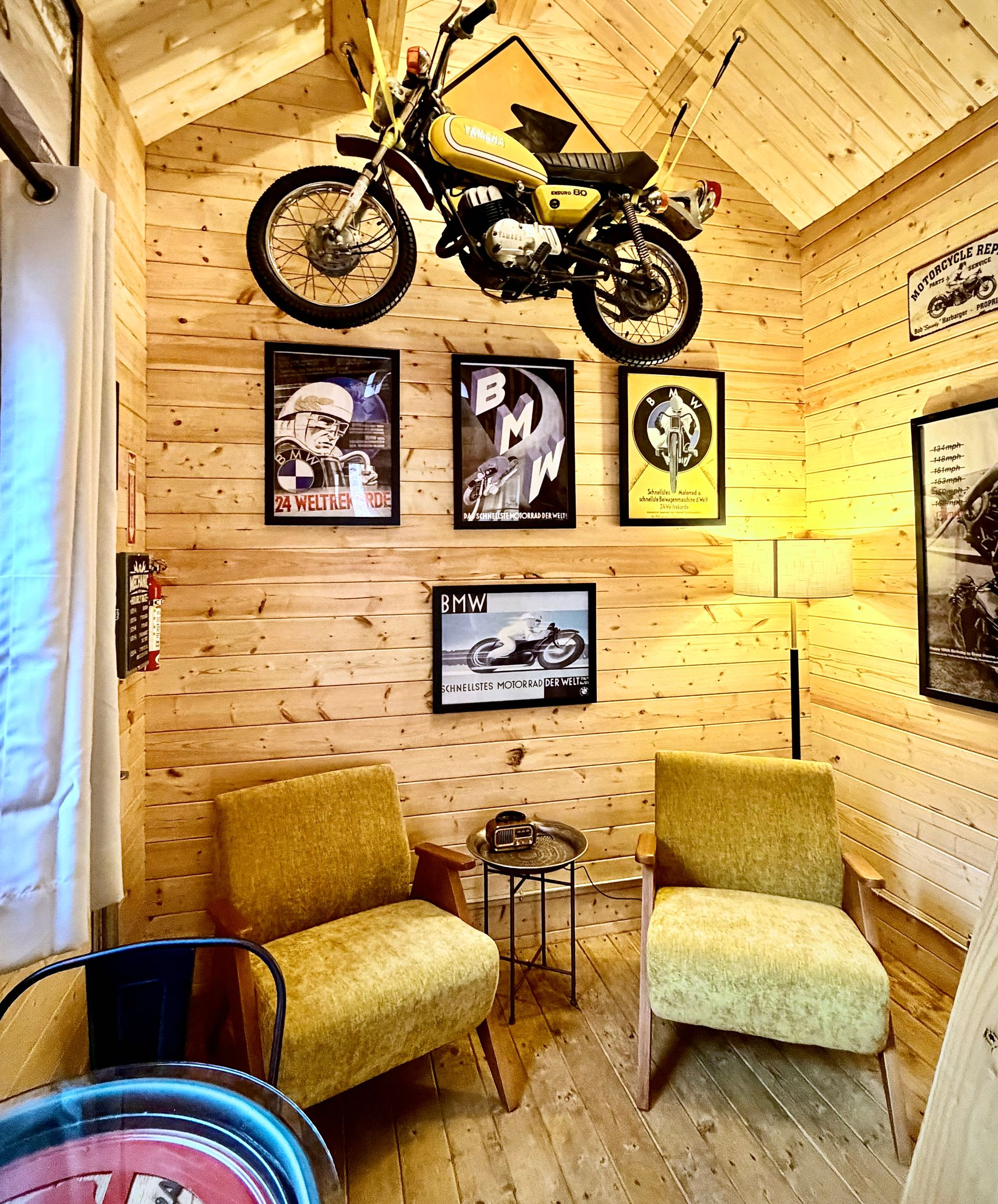 Cozy wood-paneled room with two chairs, framed art, and a motorcycle mounted high on the wall.