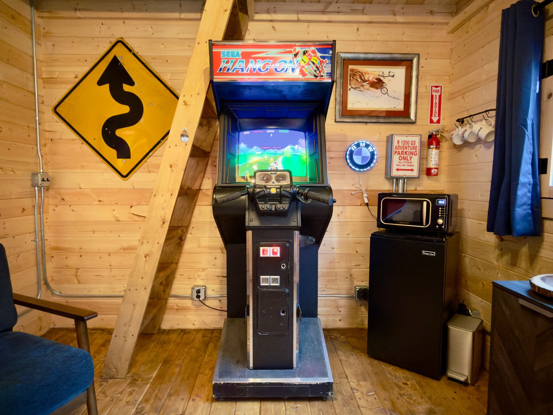 Arcade cabinet in a wooden cabin room, with a yellow winding-road sign on the wall and a small TV to the right