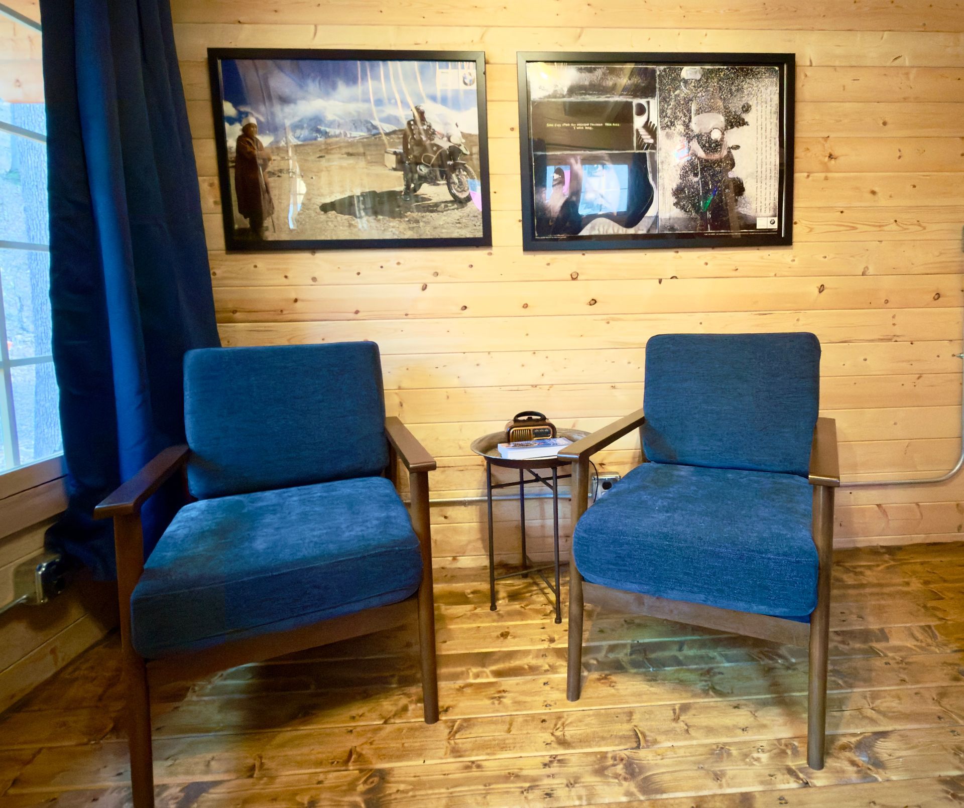 Two blue chairs beside a small table with a lamp in a wood-paneled room, with framed artwork on the wall.
