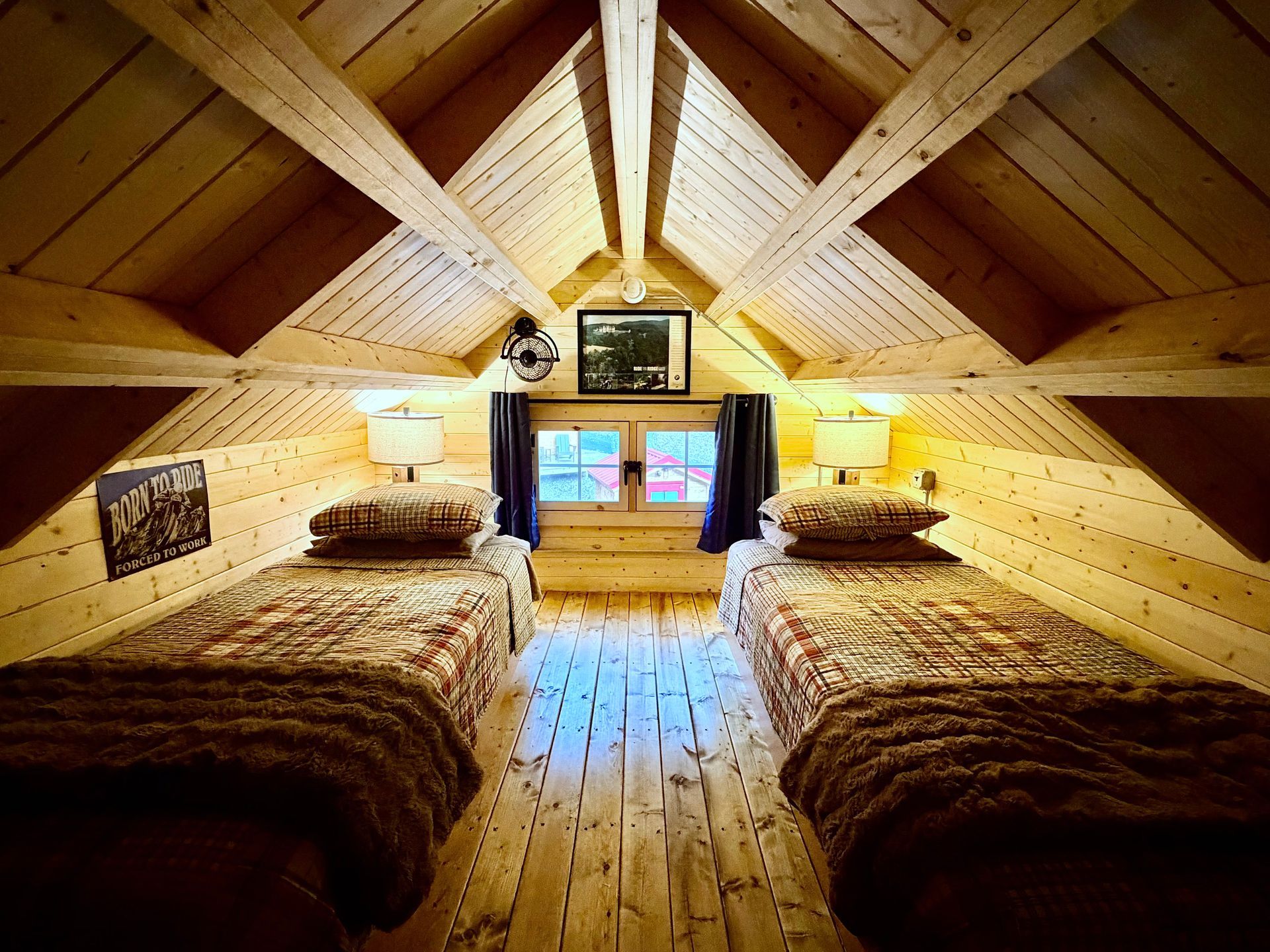 Cozy attic bedroom with two beds, wood-paneled walls, and a central window under a peaked ceiling.