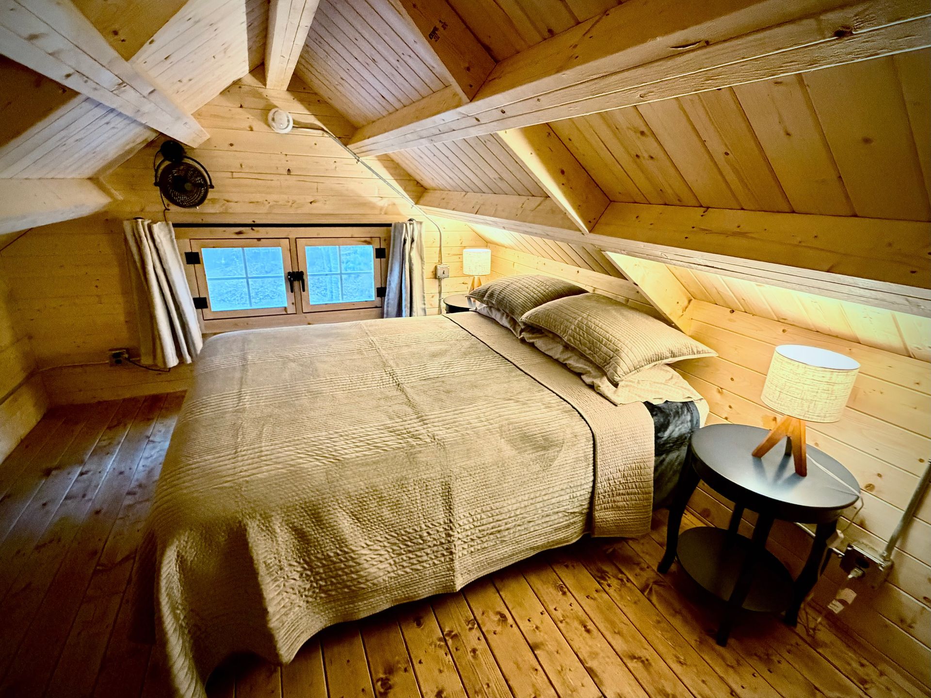 Cozy attic bedroom with sloped wood ceiling, queen bed, bedside lamp, and small windows