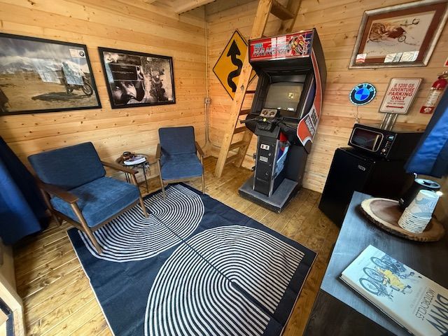 Cozy wood-paneled arcade room with two blue chairs, a patterned rug, framed art, and a vintage arcade cabinet