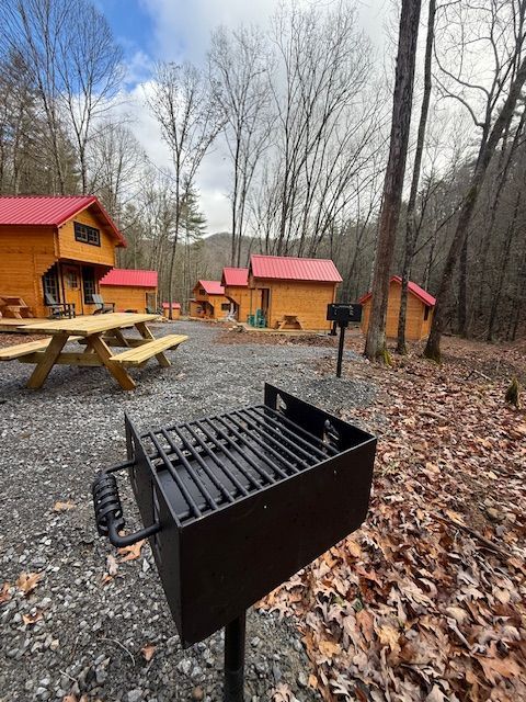 Black grill beside picnic table in a wooded campsite with small red-roof cabins.
