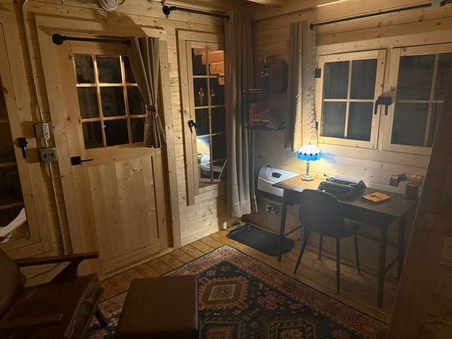 Cozy wood-paneled room with desk, blue lamp, patterned rug, and door beside two windows.