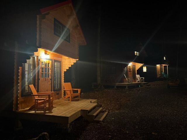 Dimly lit wooden cabins with porch lights glowing at night, one with chairs on a deck, in a gravel lot