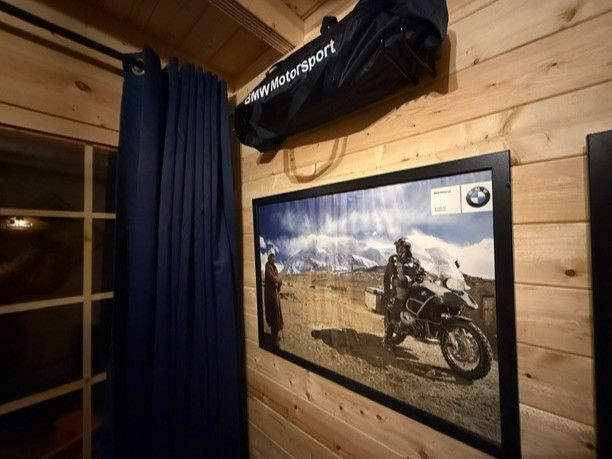 Cozy wood-paneled room with blue curtains, framed motorcycle photo, and a hanging black duffel bag.