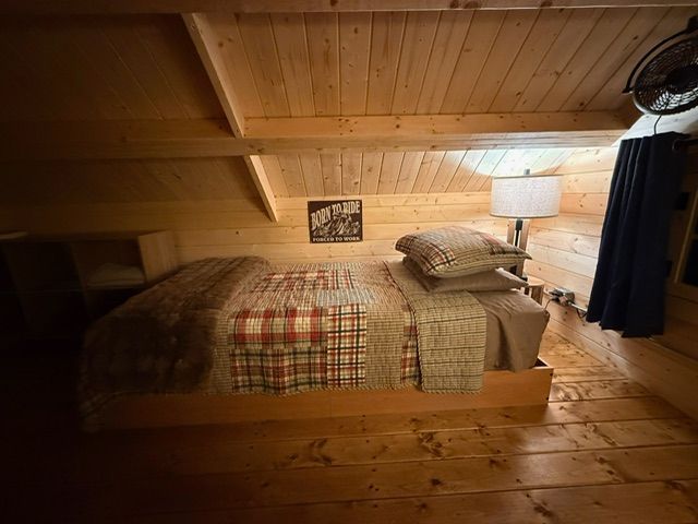 Cozy loft bedroom with a wooden bed, plaid quilt, lamp, and sloped wood-paneled ceiling
