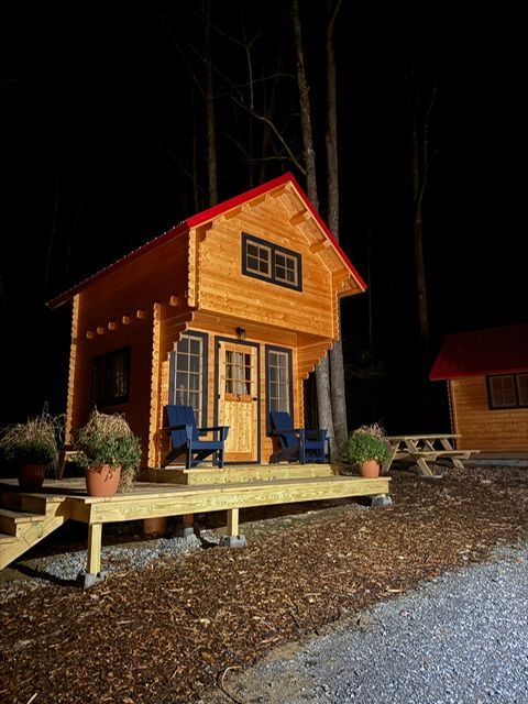 Small wooden cabin with blue chairs on a lit porch at night, surrounded by gravel and trees.