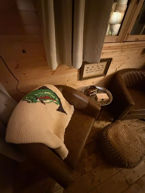 Cozy cabin corner with chair, blanket, woven hat, and small side table by a curtained window.