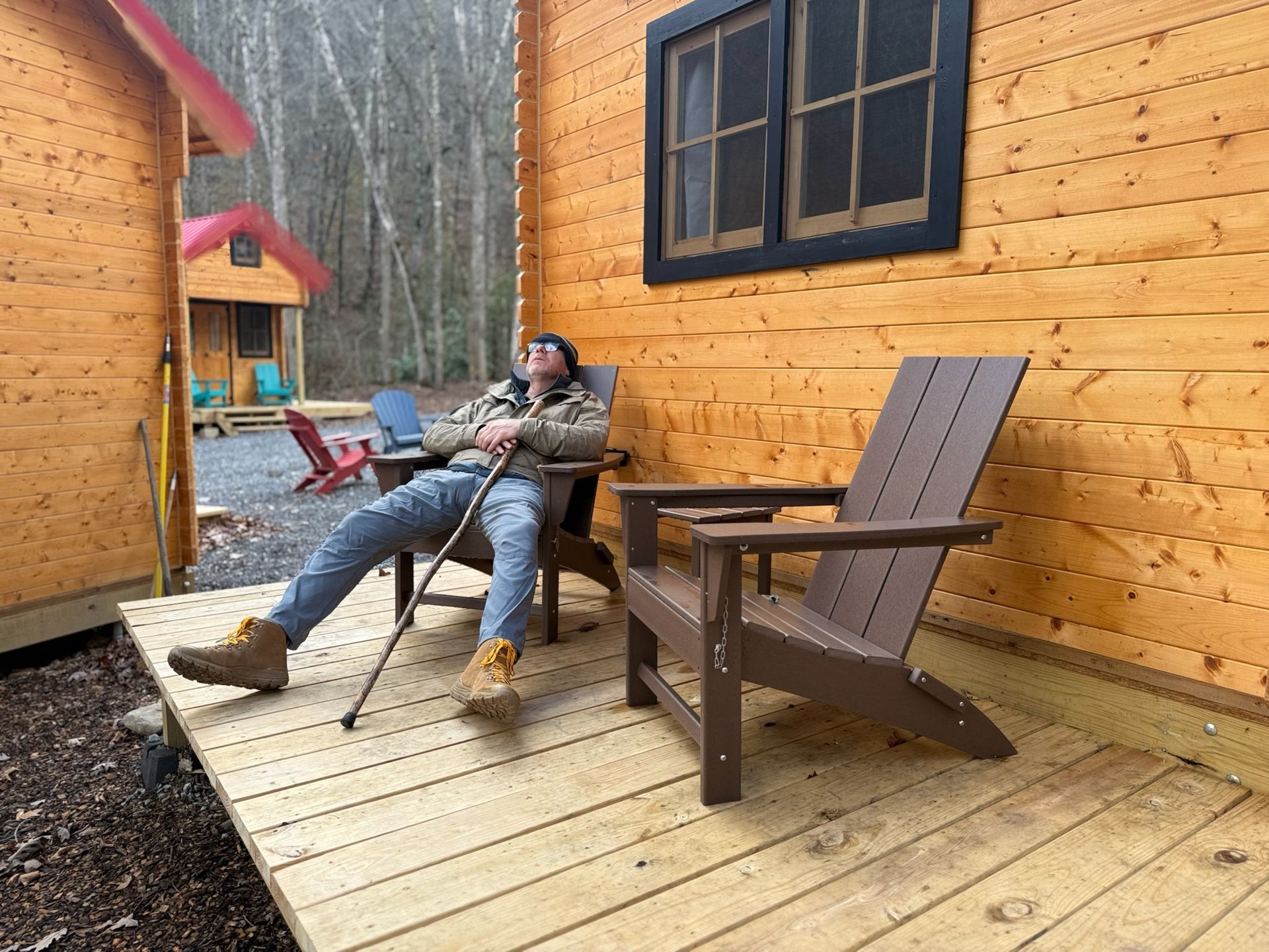 Person relaxing in a chair on a wooden cabin porch with a table and Adirondack chairs nearby.