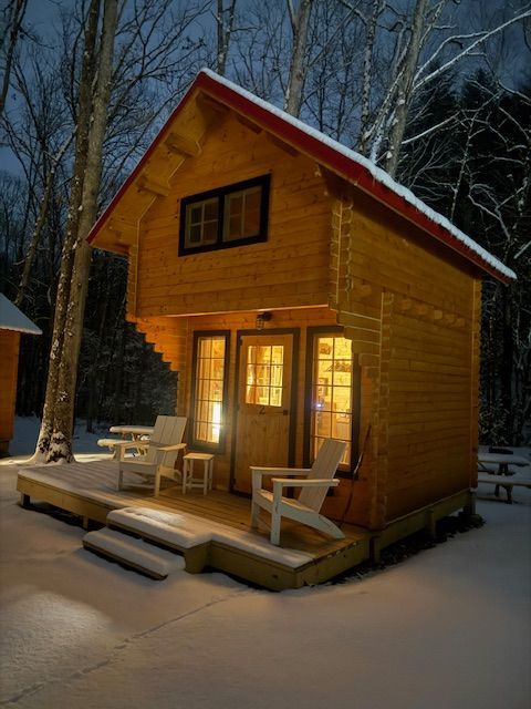 Small wooden cabin glowing warmly at night in a snowy forest, with a porch and chairs.