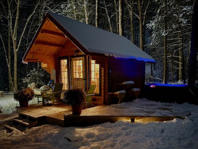 Snowy cabin lit warmly at night, with a porch, deck chairs, and a forest backdrop