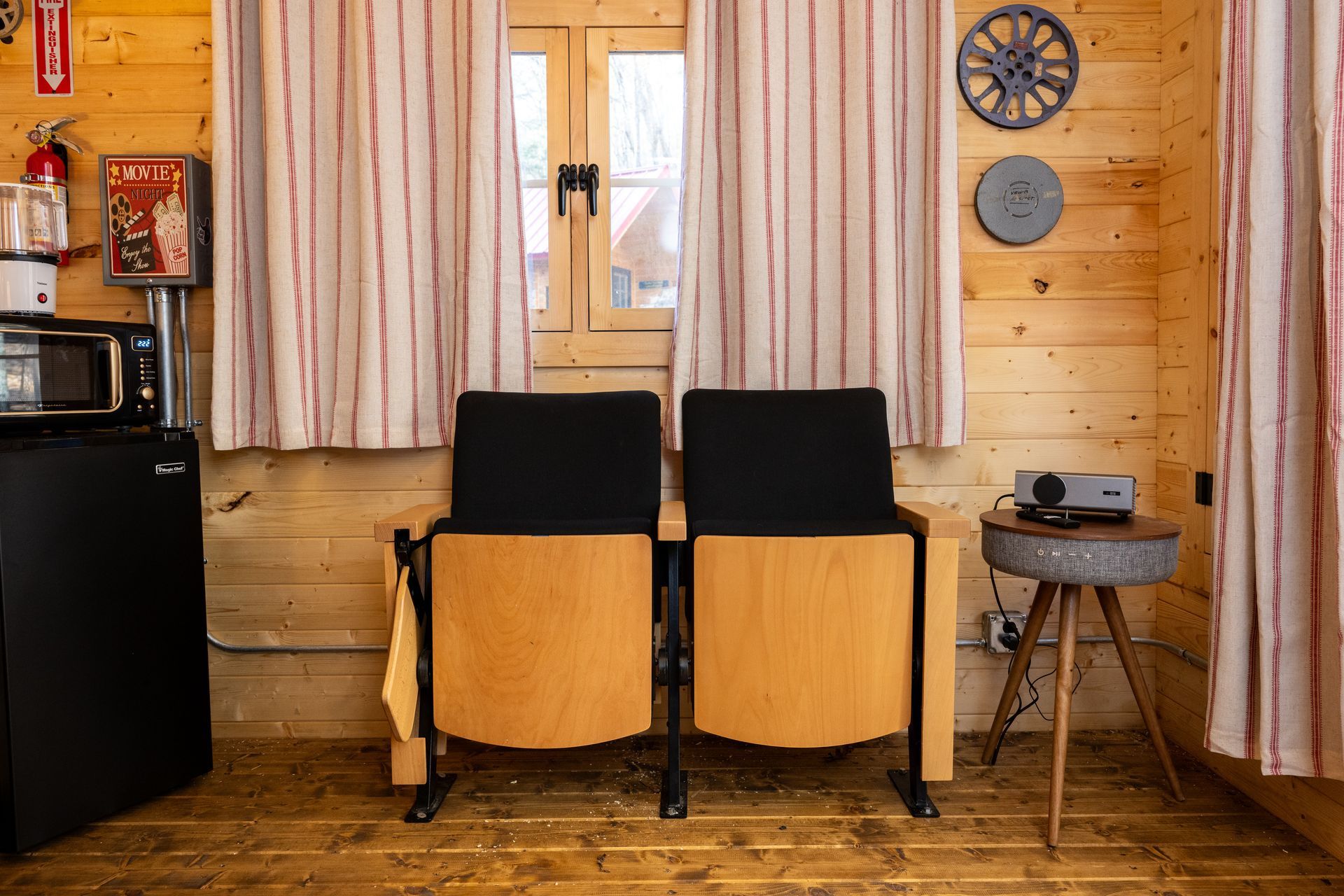 Cozy wooden cabin interior with two black chairs, window, small table, curtains, and a mini fridge