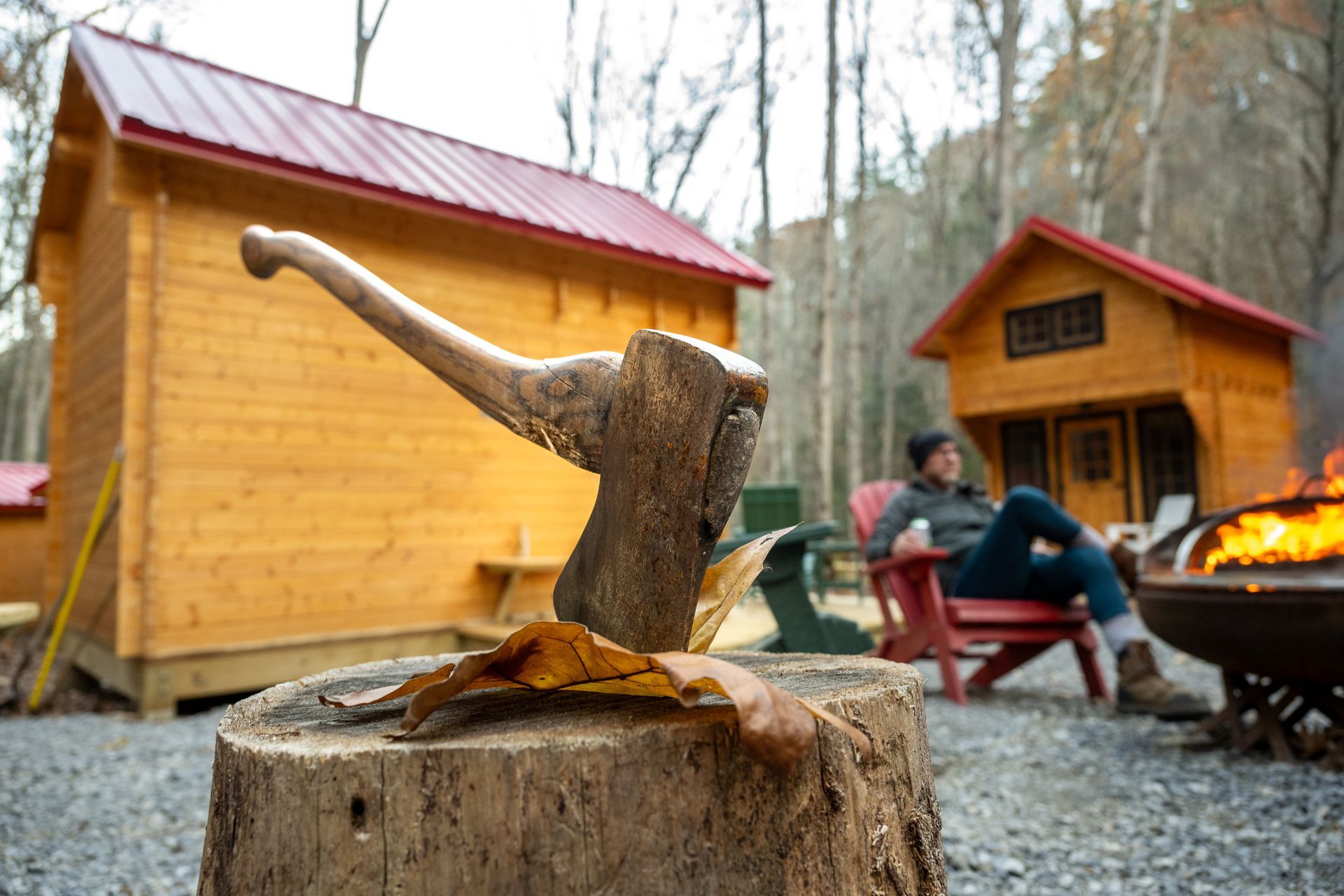 Axe embedded in a chopping block near cabins and a fire pit, with a person sitting by the flames outdoors.