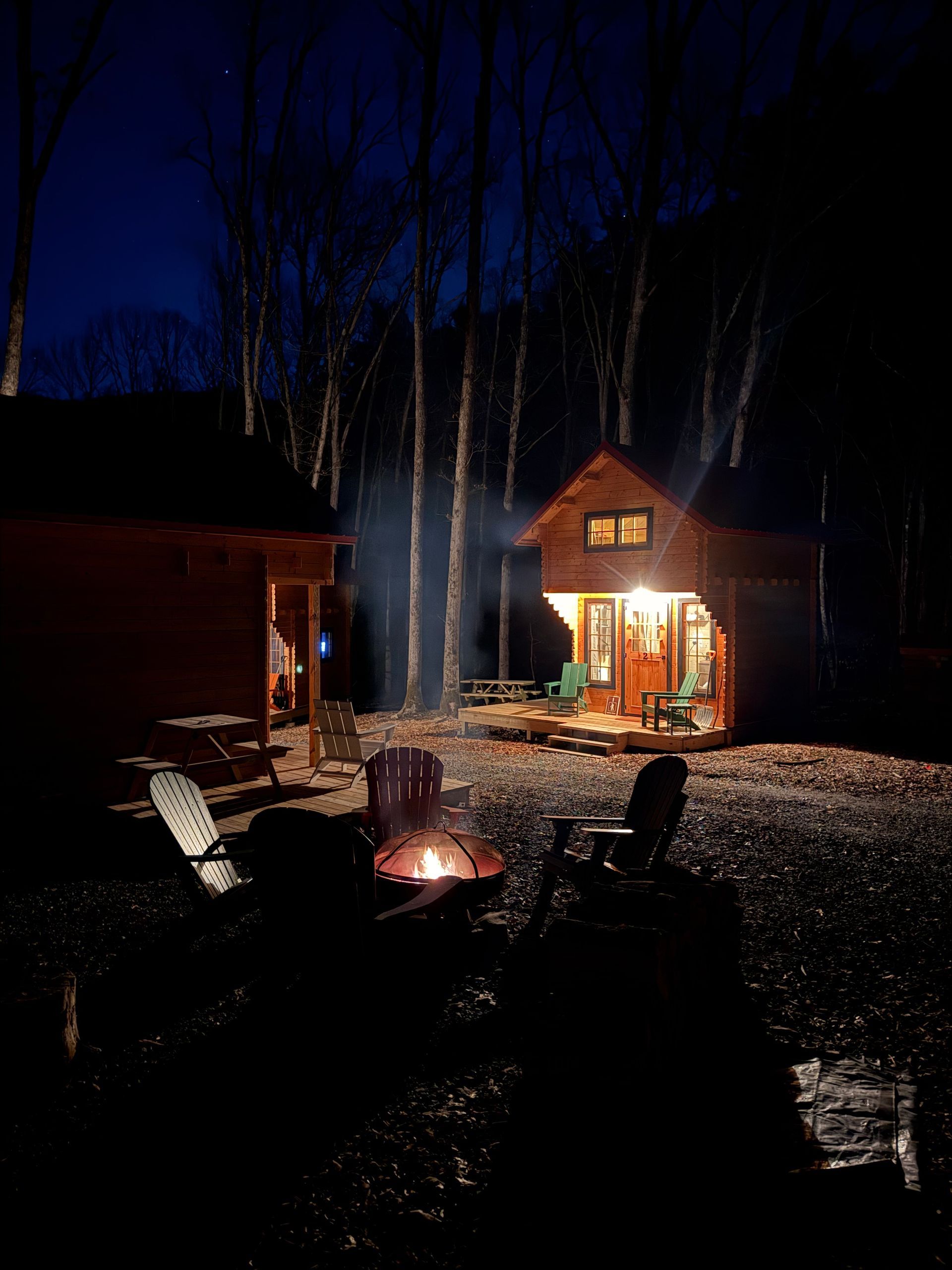 Nighttime cabin yard with a glowing fire pit, Adirondack chairs, and a lit house under a dark blue sky
