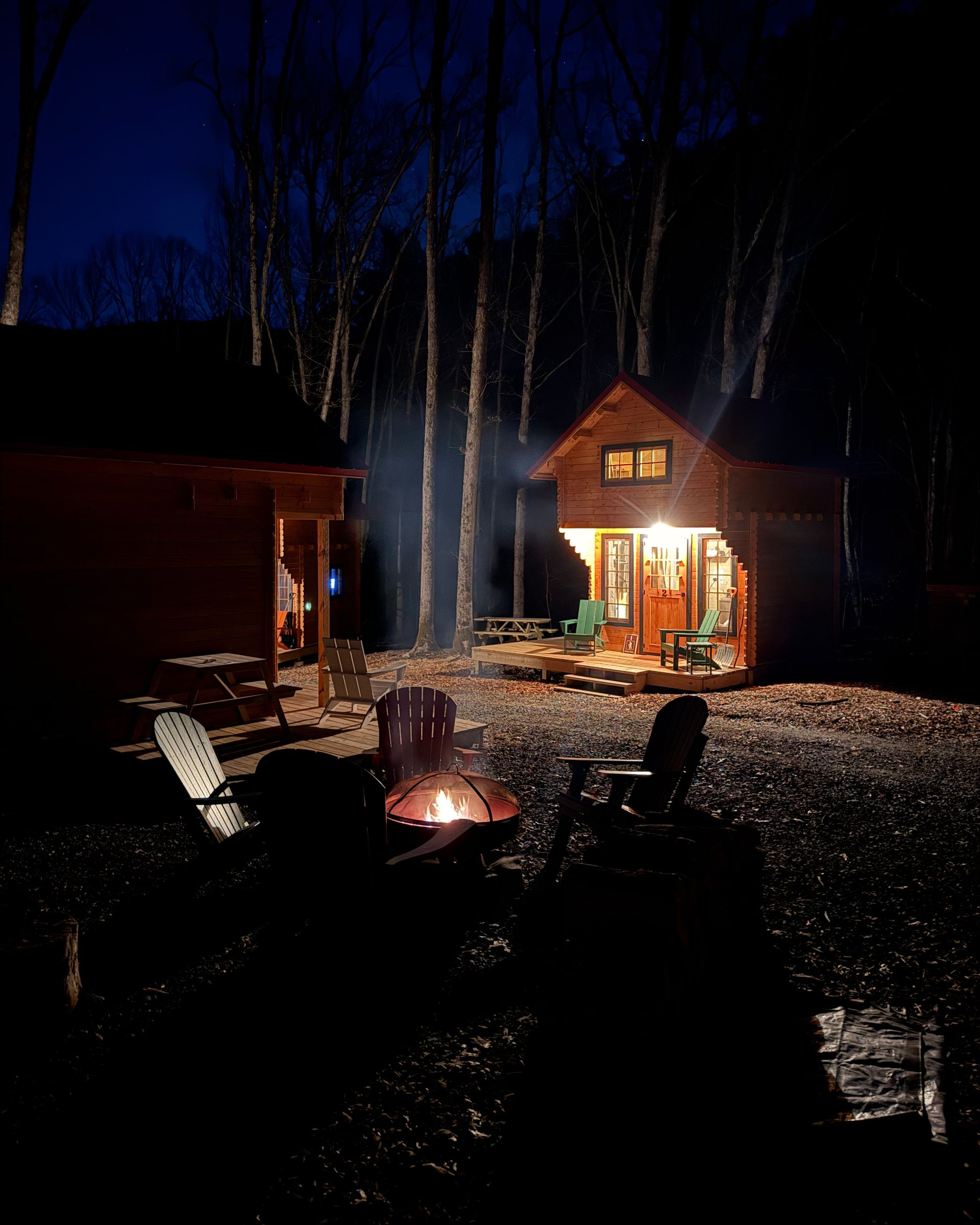 Nighttime cabin scene with a glowing fire pit and lit cottage in a snowy forest clearing