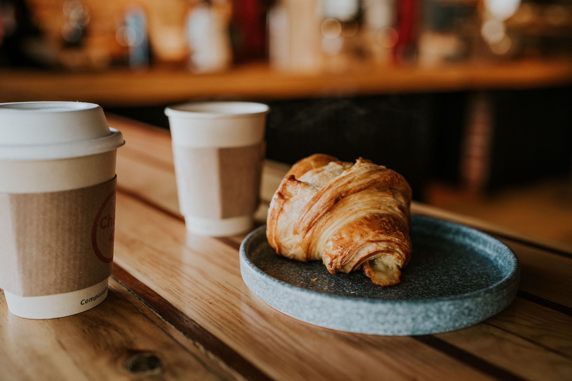 Coffee cup and croissant on a wooden café table with a blurred warm background