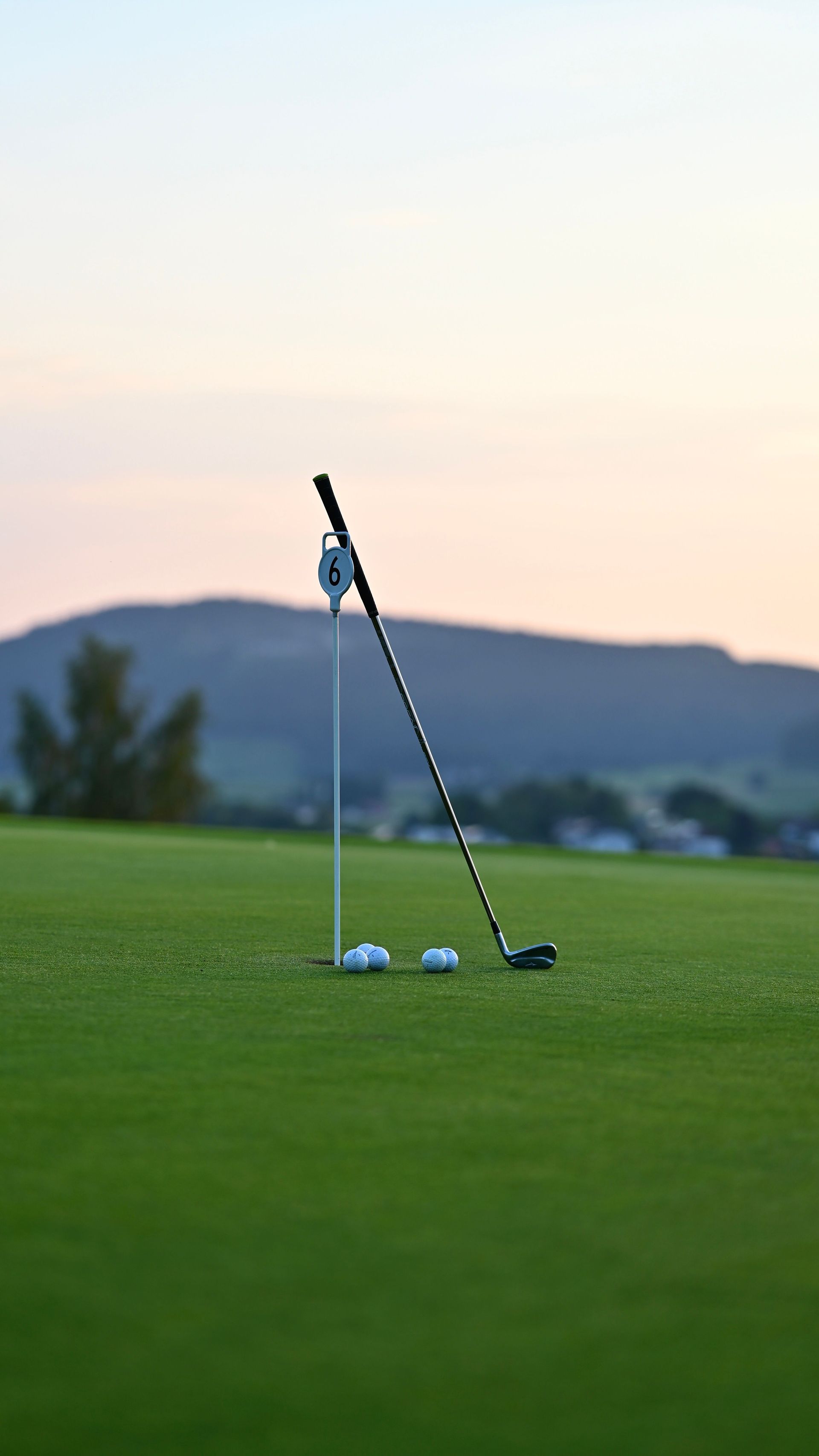 Golf club leaning on a green putting surface at sunset, with two golf balls and a distant hill backdrop