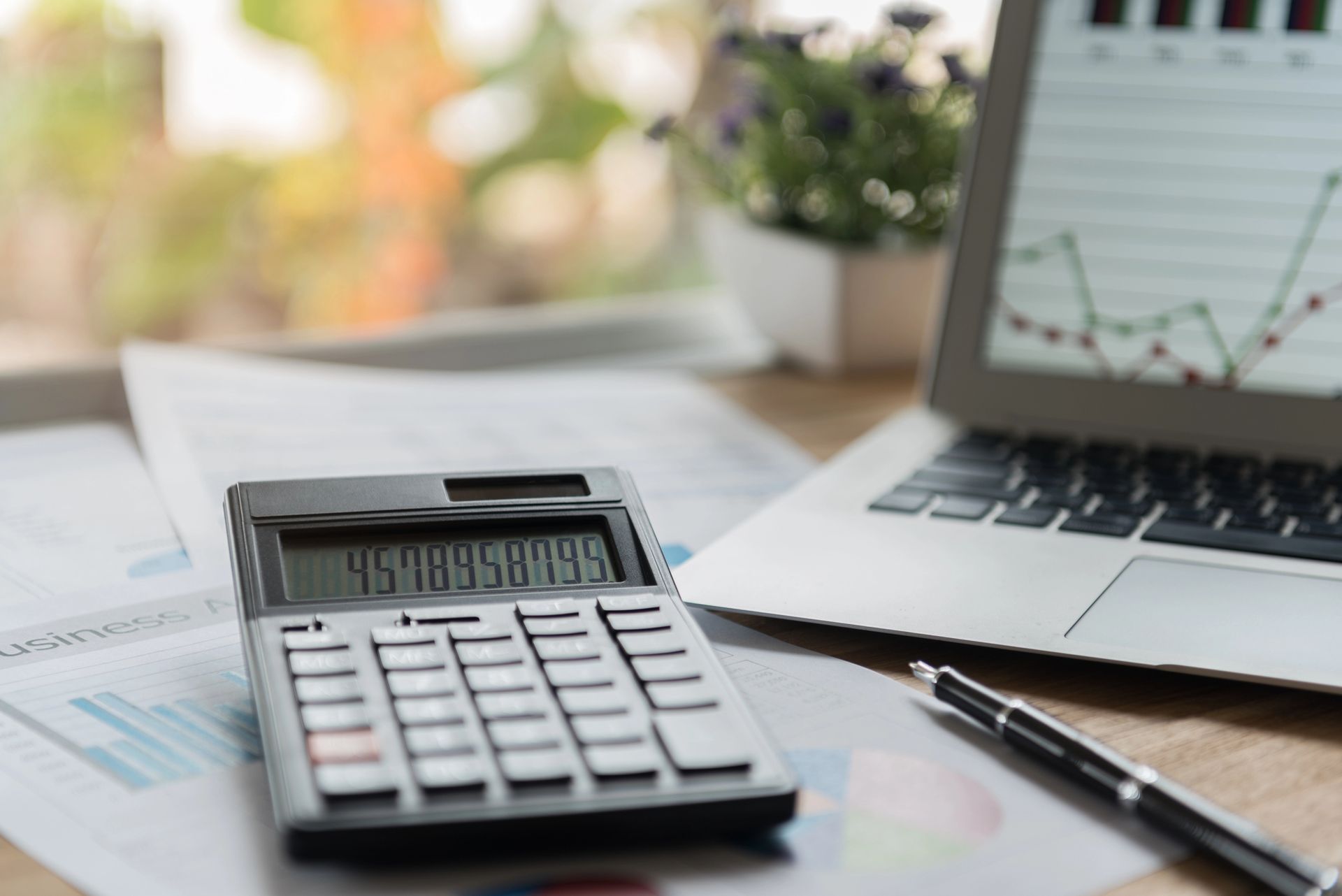 Calcolatrice, computer portatile e penna su una scrivania con grafici finanziari sullo sfondo.