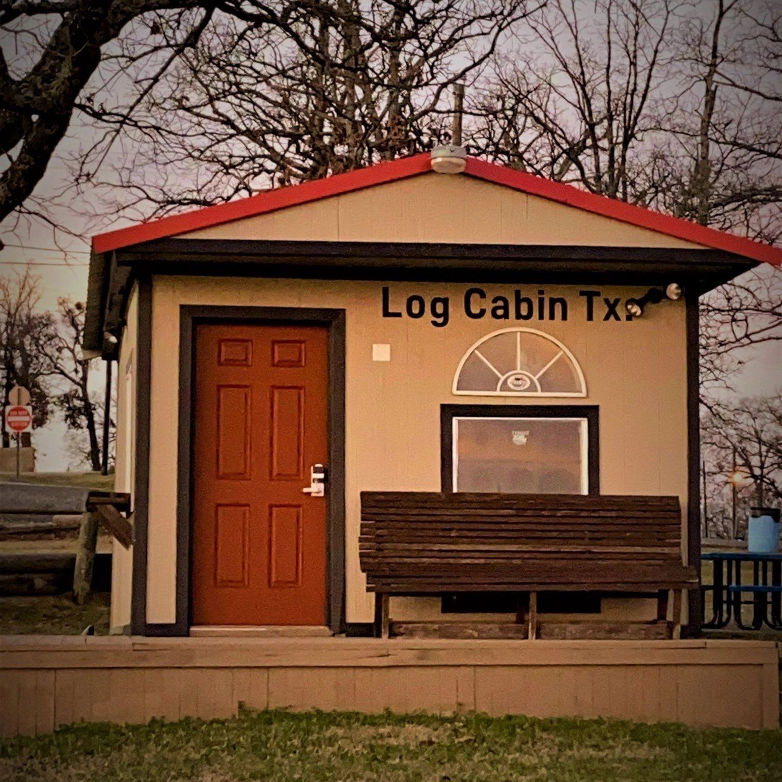 Small tan log cabin building with red roof, wooden door, bench, and “Log Cabin Tx” sign.