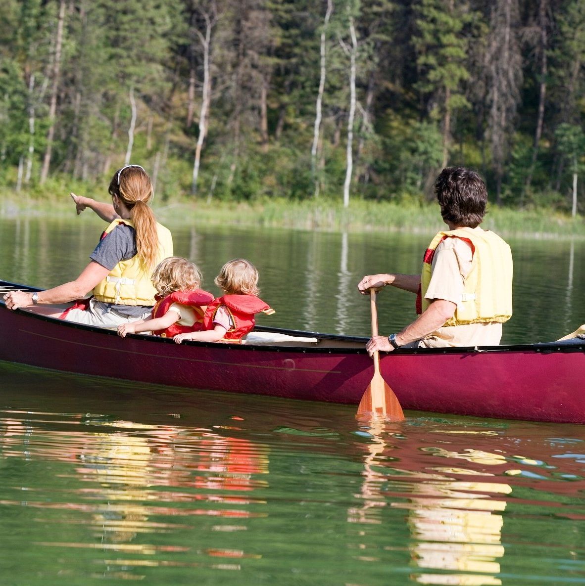 Three people canoeing on a calm green lake with a forested shoreline in the background.