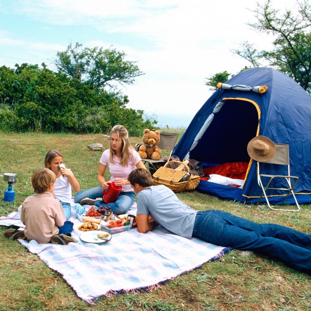 Friends picnicking on a blanket beside a blue tent in a grassy campsite