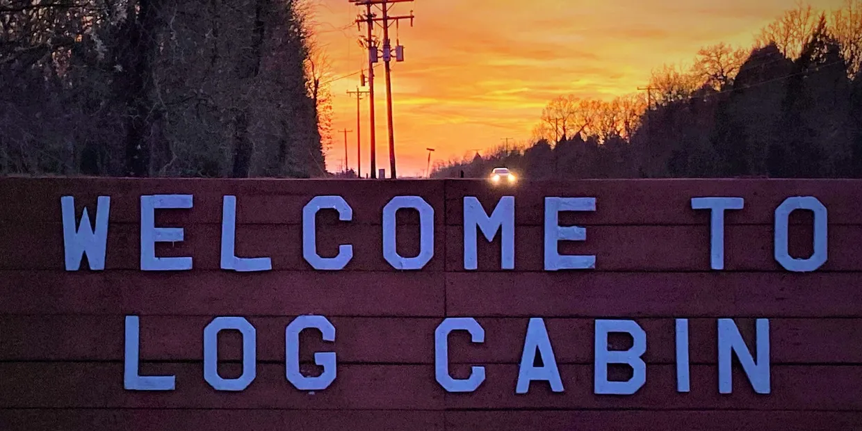 Welcome to Log Cabin sign in purple letters at sunset, with trees and utility poles behind it