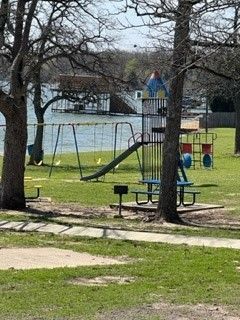 Empty playground with slide, picnic table, and trees near a waterfront on a grassy day