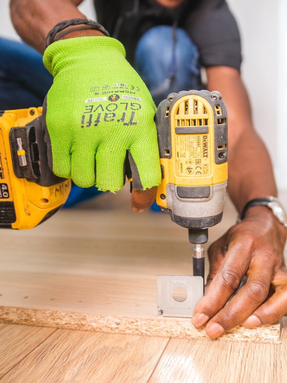 Gloved hands using a yellow drill to fasten a metal bracket to a wooden board