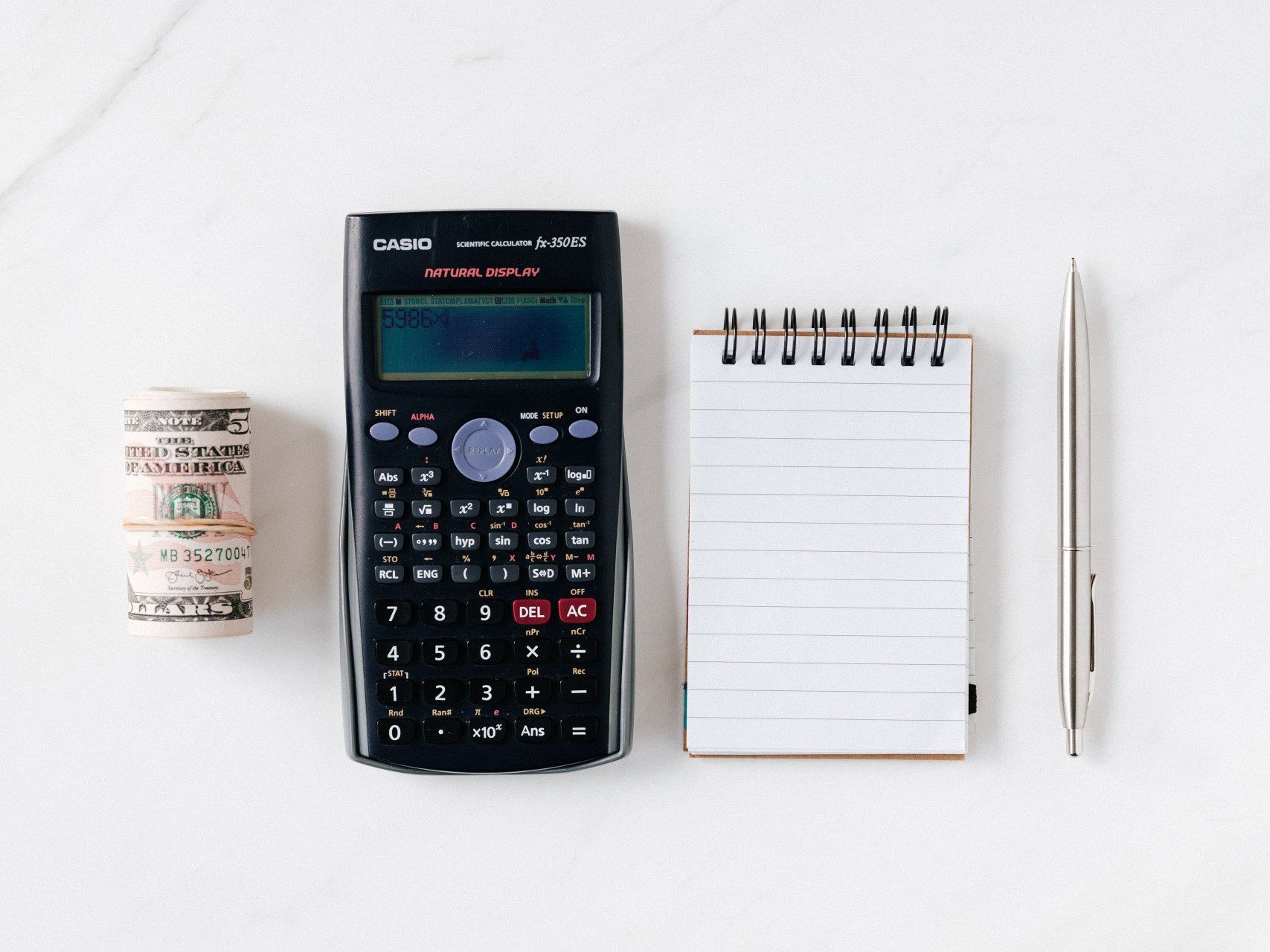 Calculator, notepad, pen, and small roll of tape arranged neatly on a white desk