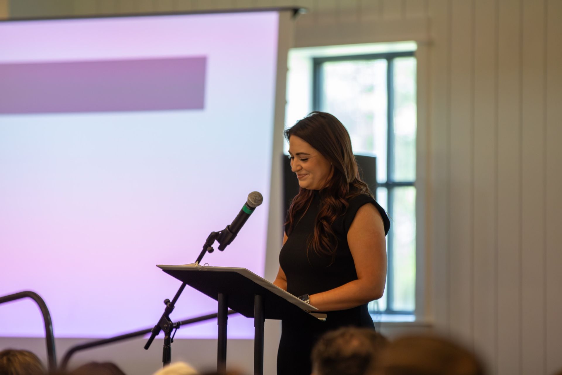 Woman speaking at a podium with a microphone in a conference room, with a presentation screen behind her.