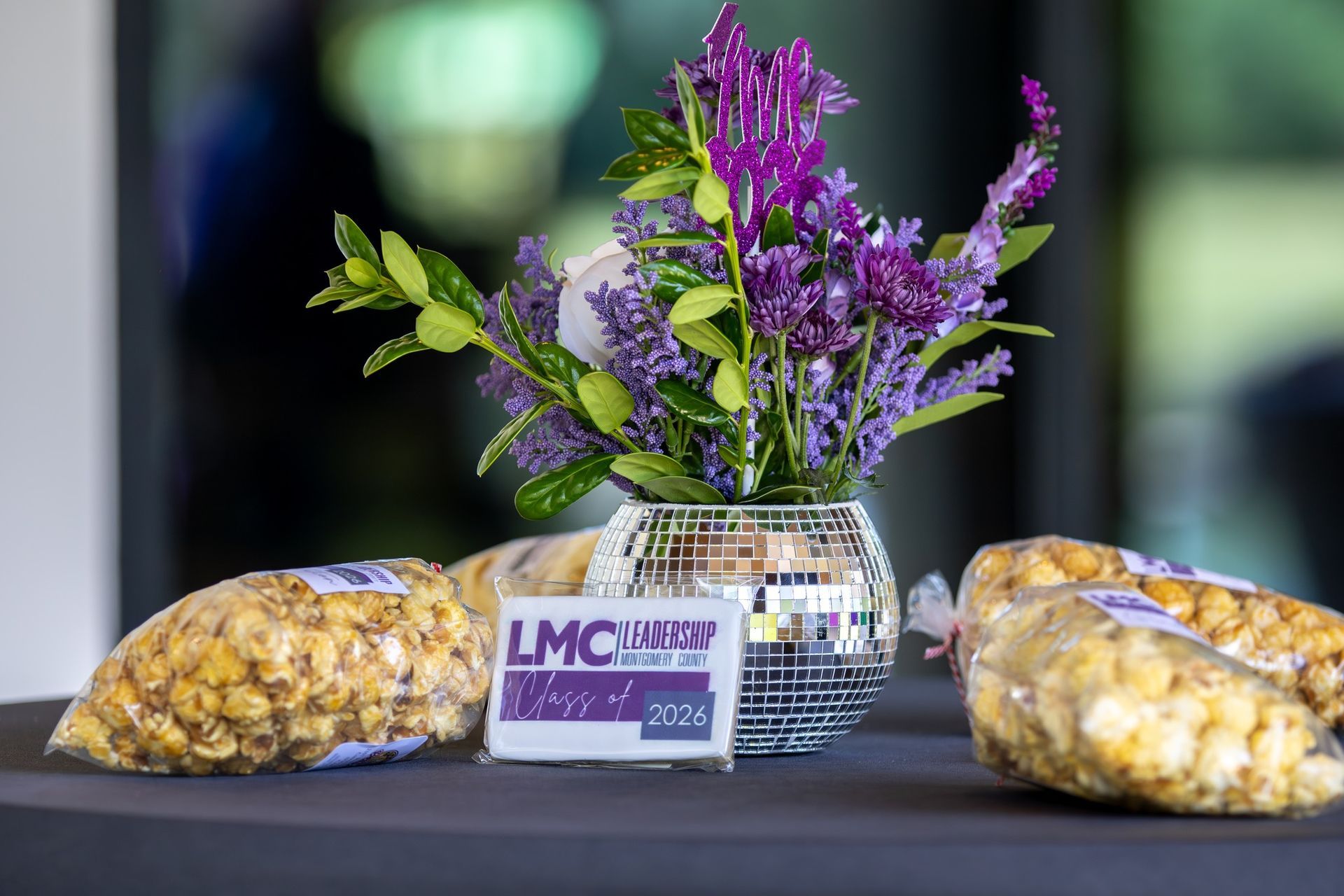 Purple floral centerpiece with an LMC sign between two bags of snacks on a table