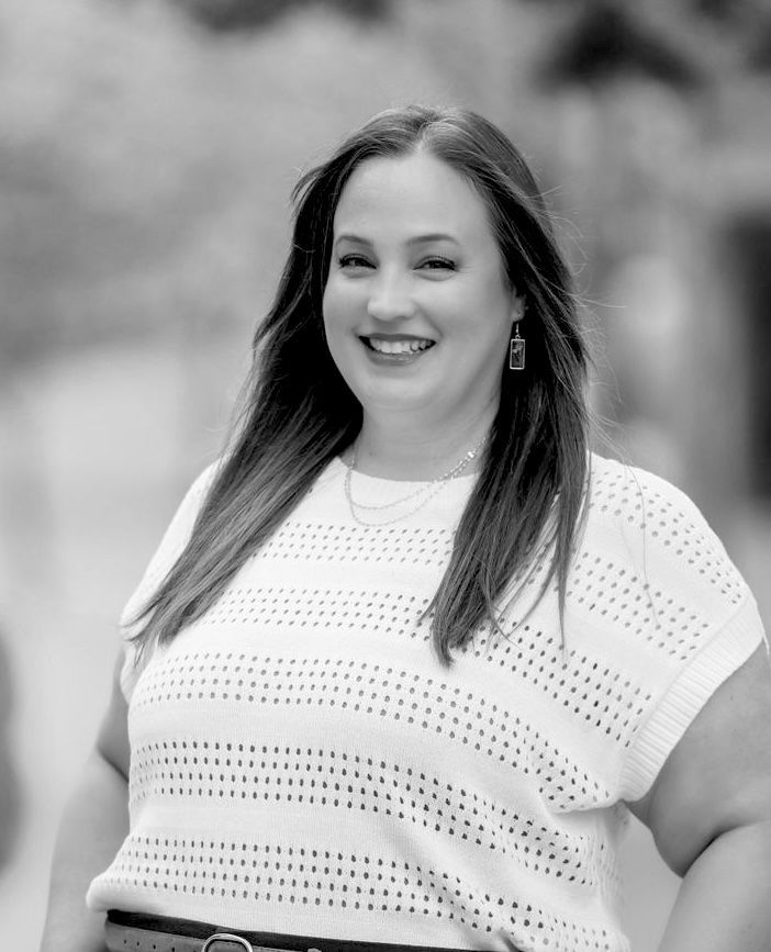 Smiling woman in a white knit top standing outdoors, with blurred trees in the background