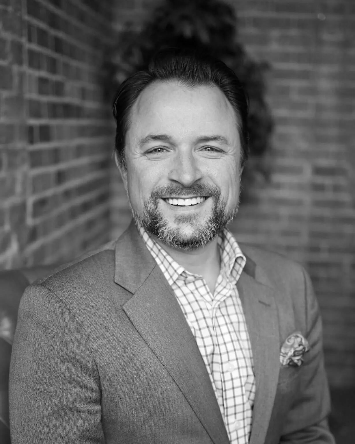 Smiling man in a blazer and checkered shirt posing in front of a brick wall.