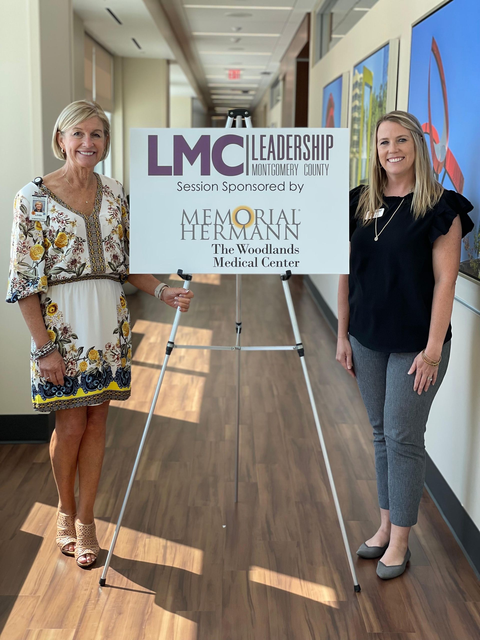 Two women hold a LMC Leadership sign in a bright hallway with flags and sunlight.
