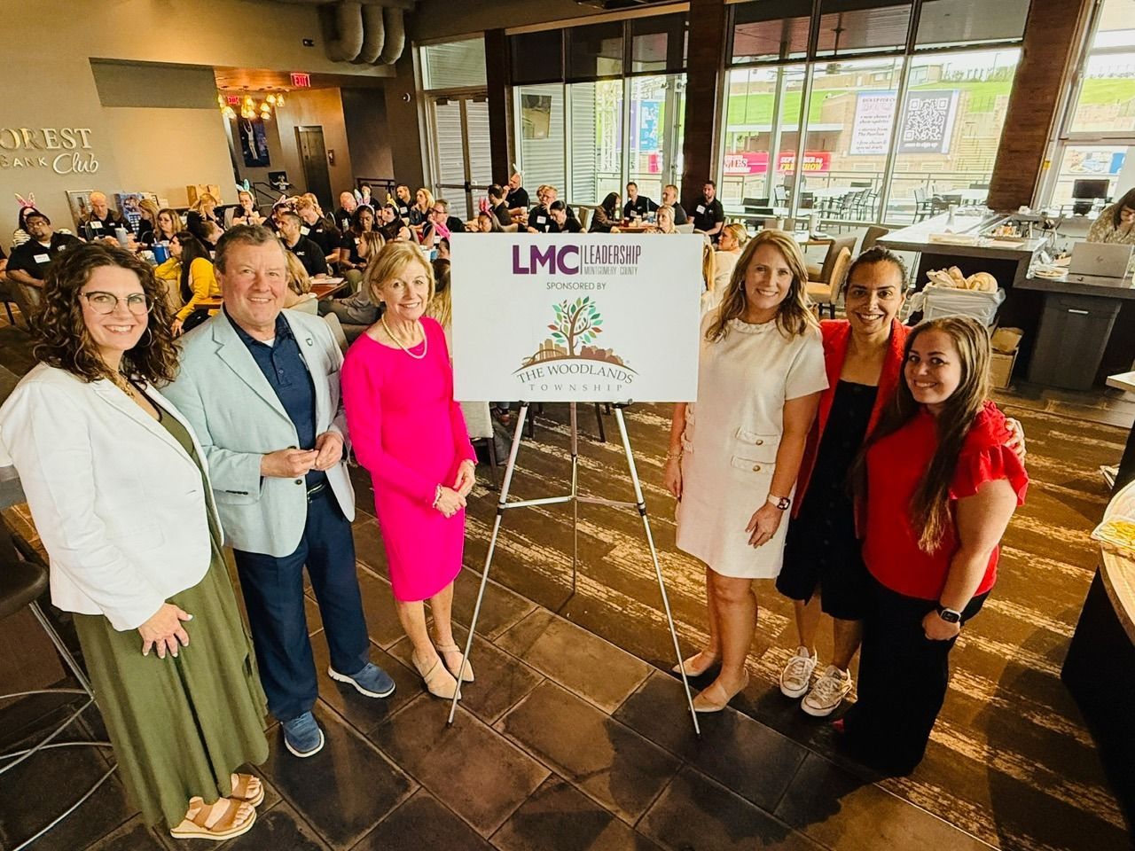 Group standing by an LMC sign at an indoor event, with tables, chairs, and guests in the background