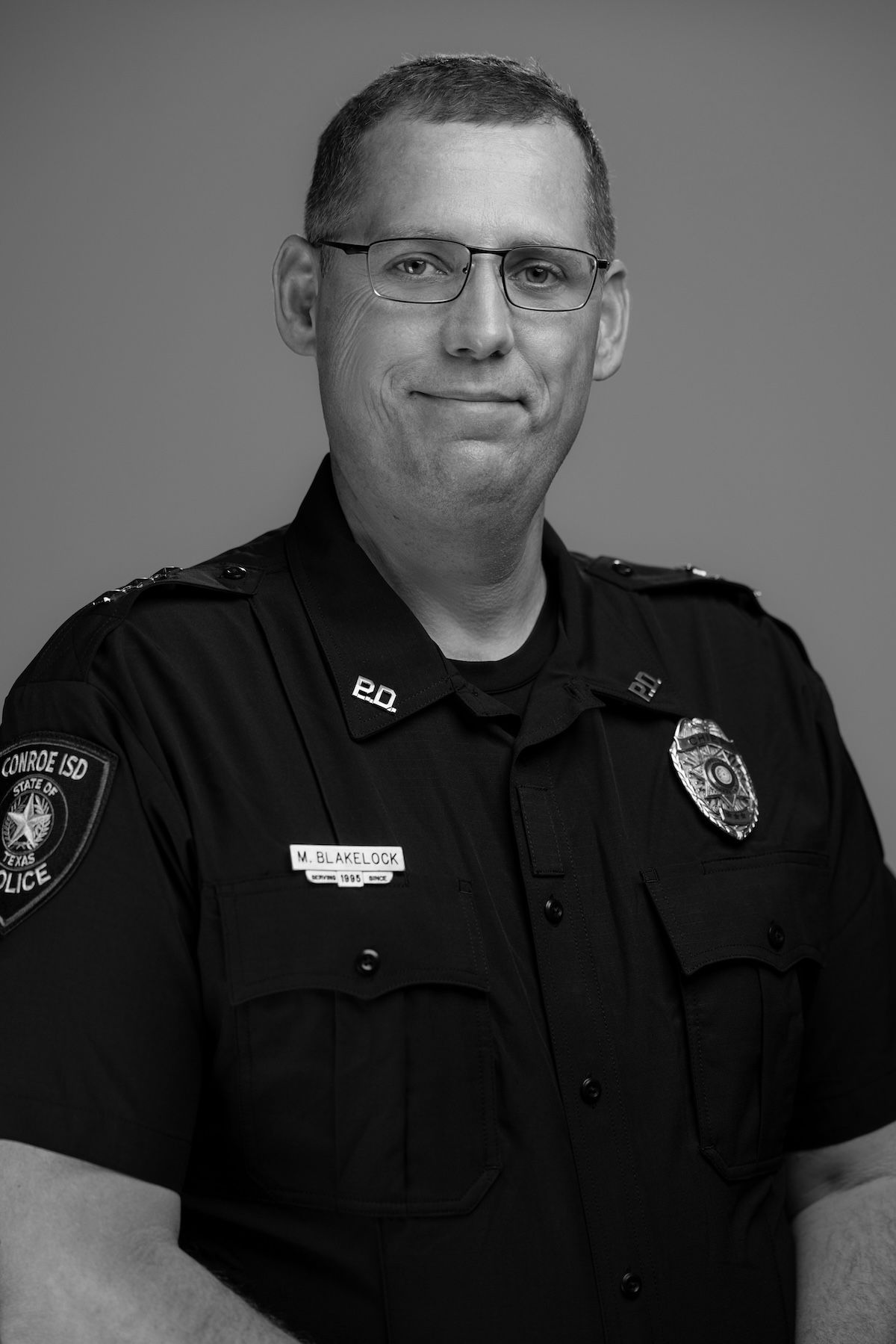 Police officer in dark uniform and glasses, standing against a plain background.