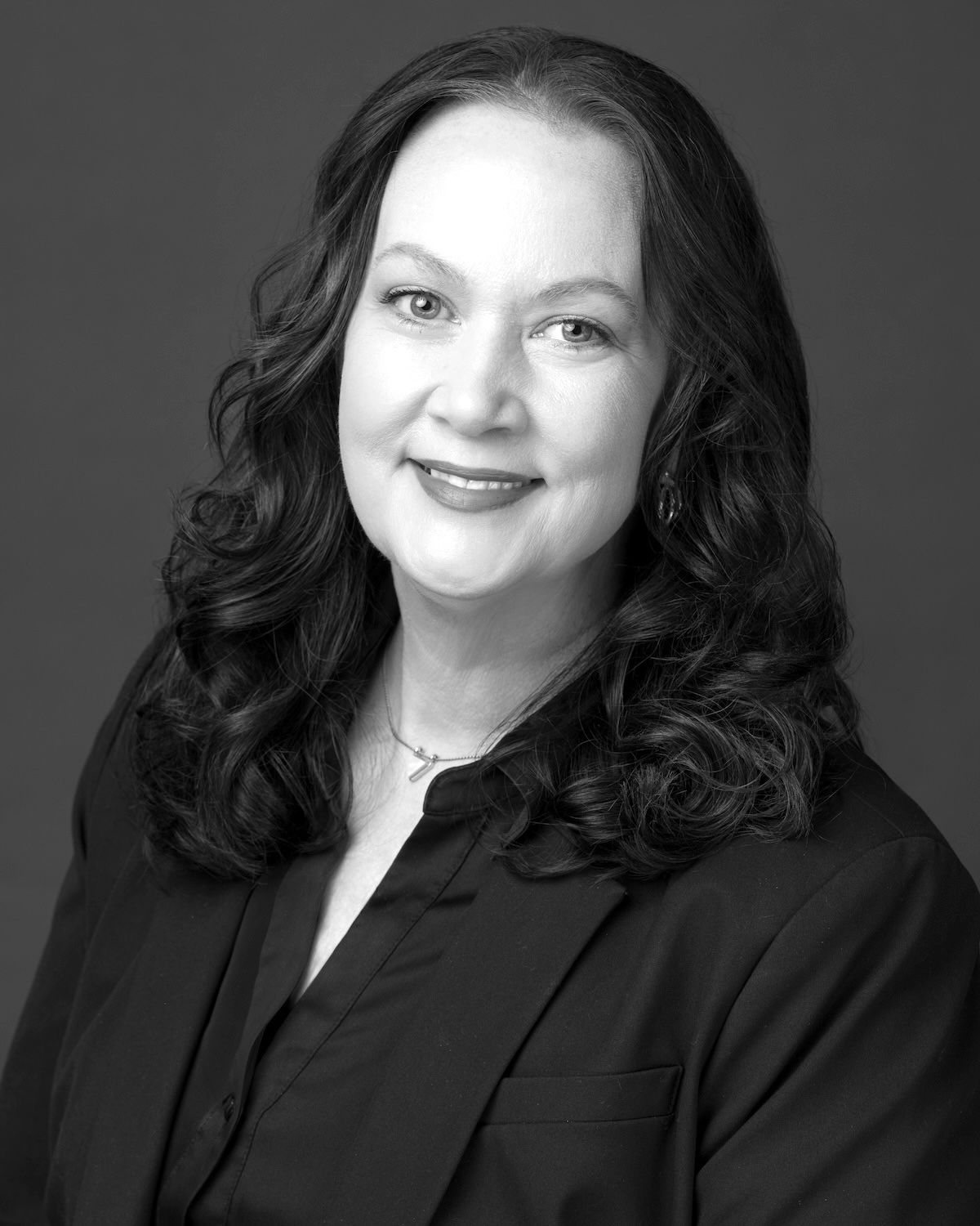 Black-and-white studio headshot of a smiling person with wavy hair in a dark blazer.
