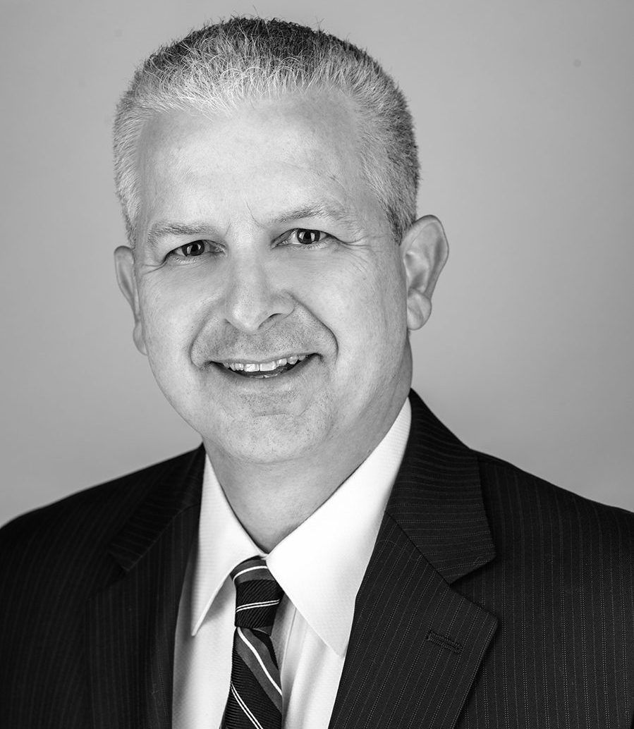 Smiling man in a dark suit and striped tie against a plain gray background