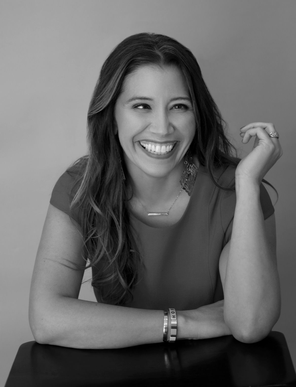 Smiling person leaning on a table, hand raised, in a black-and-white portrait.