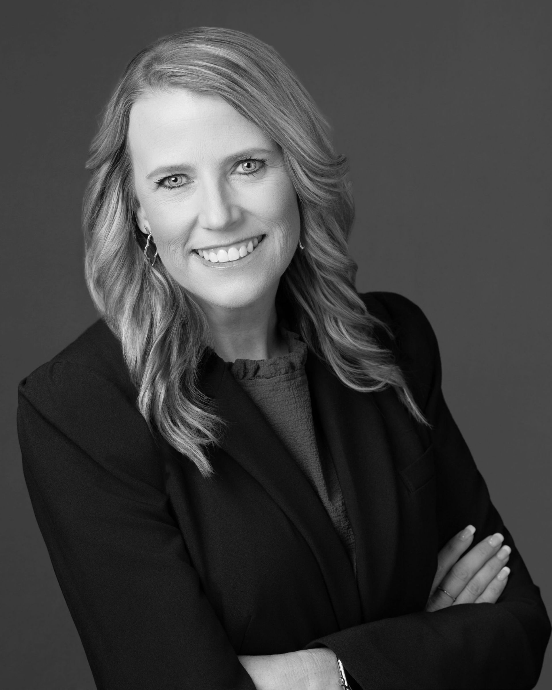 Smiling person in a dark blazer with arms crossed against a plain gray background
