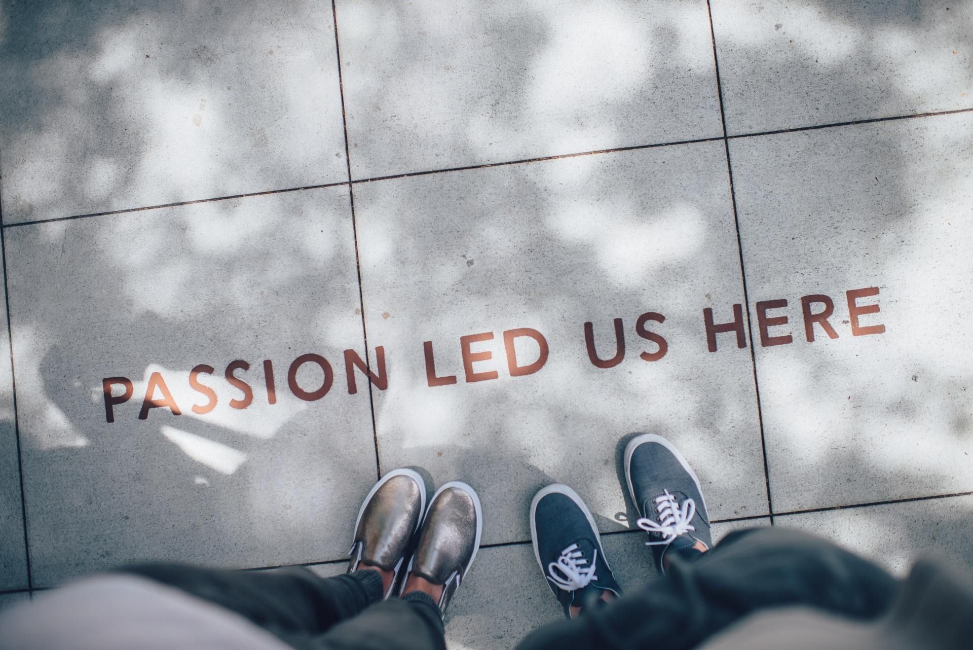 Two pairs of shoes on pavement beside red text reading “PASSION LED US HERE”