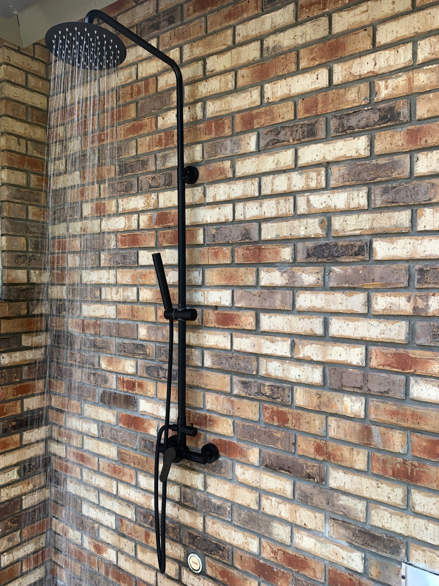 Outdoor black shower against a brick wall, with water running from the showerhead.