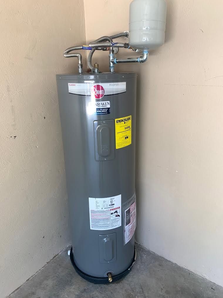 Gray water heater tank in a corner, with pipes and a small white expansion tank above it.