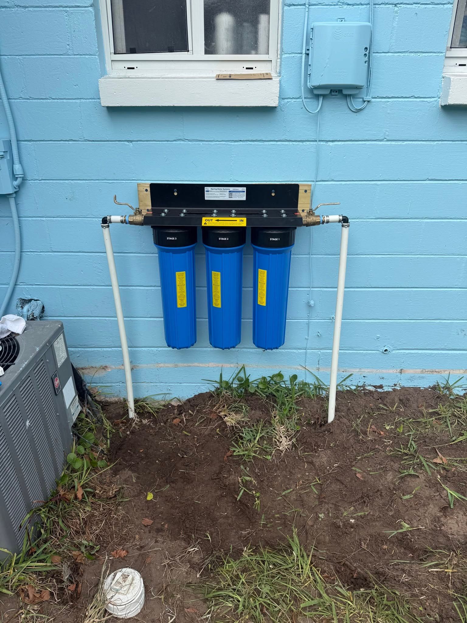 Three blue water filter housings mounted on a wall under a window outdoors.