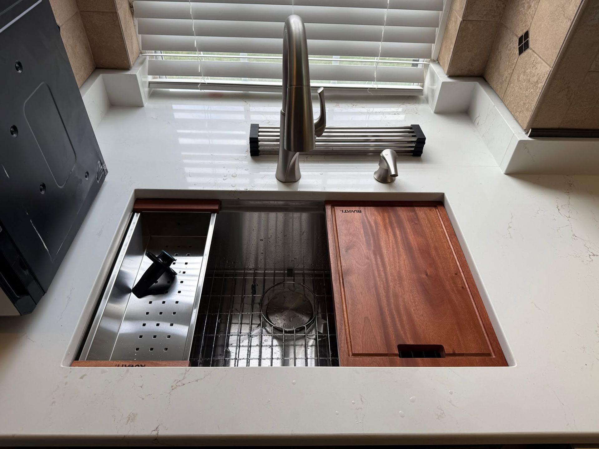 Kitchen sink with stainless basin, chrome faucet, and wooden cutting board beside a window.