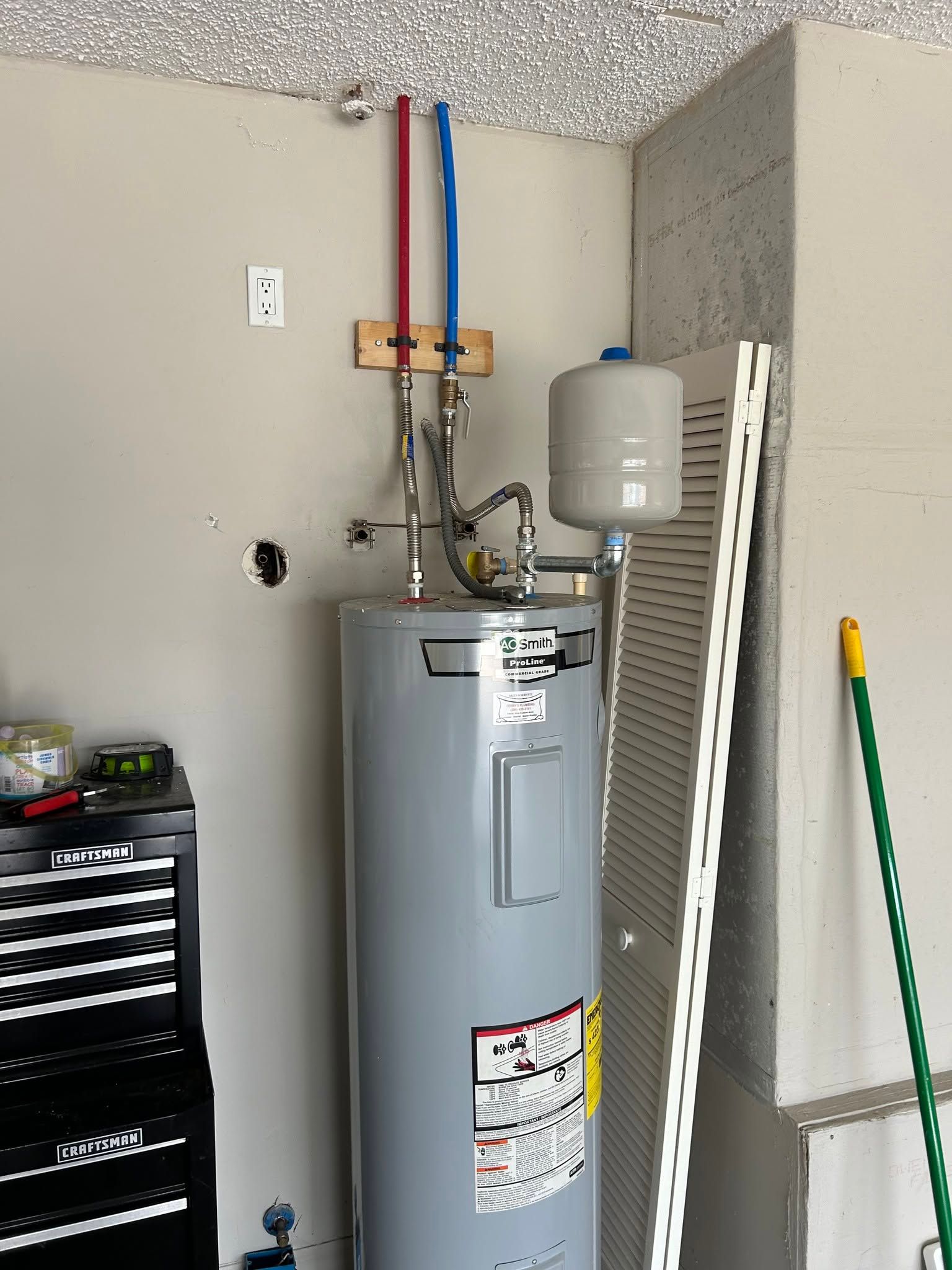 Water heater and plumbing pipes in a garage or utility room, with cleaning tools and a tool chest nearby.