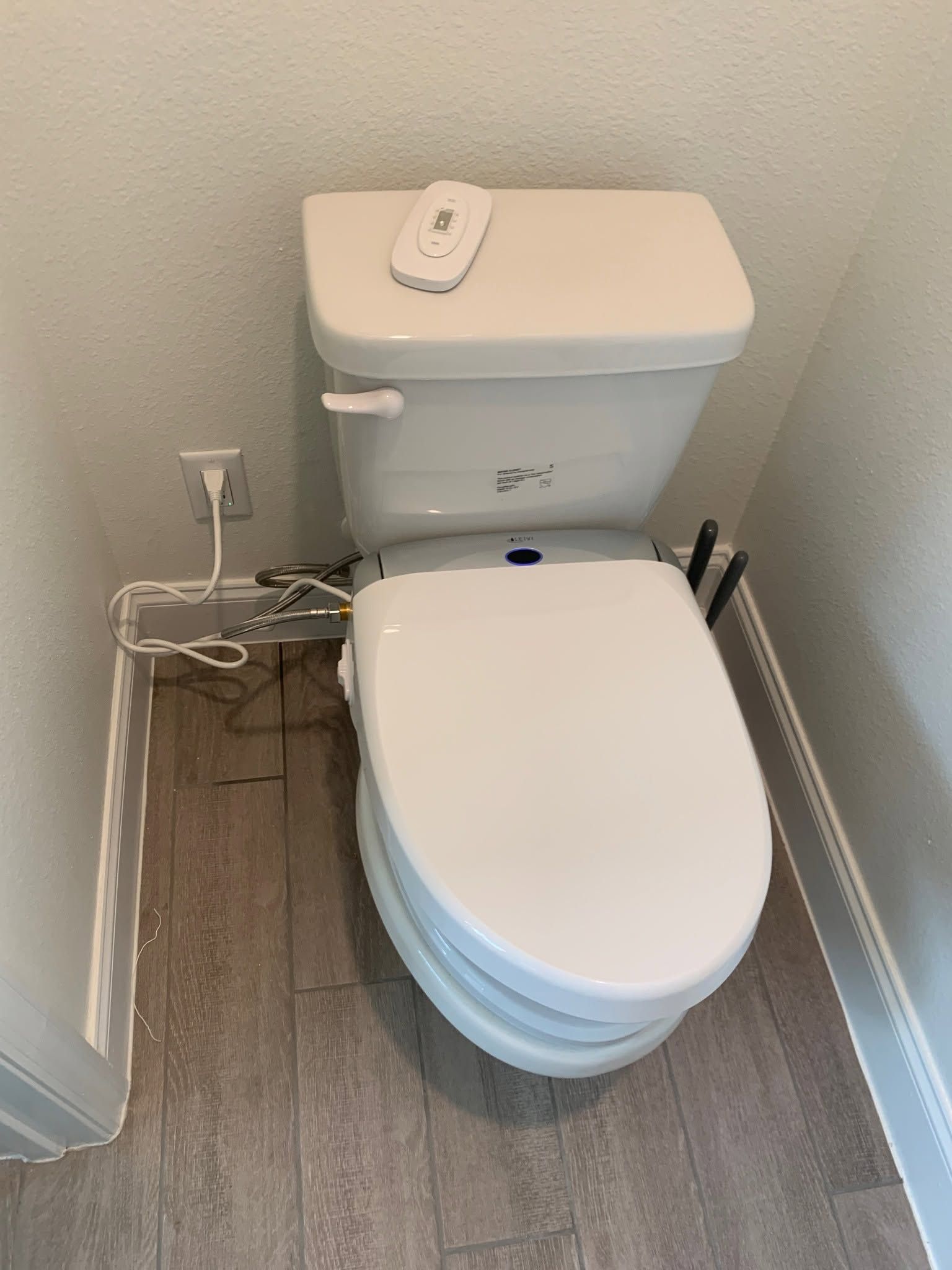 Compact bathroom with white toilet, remote on tank, and visible outlet and cords beside gray flooring.