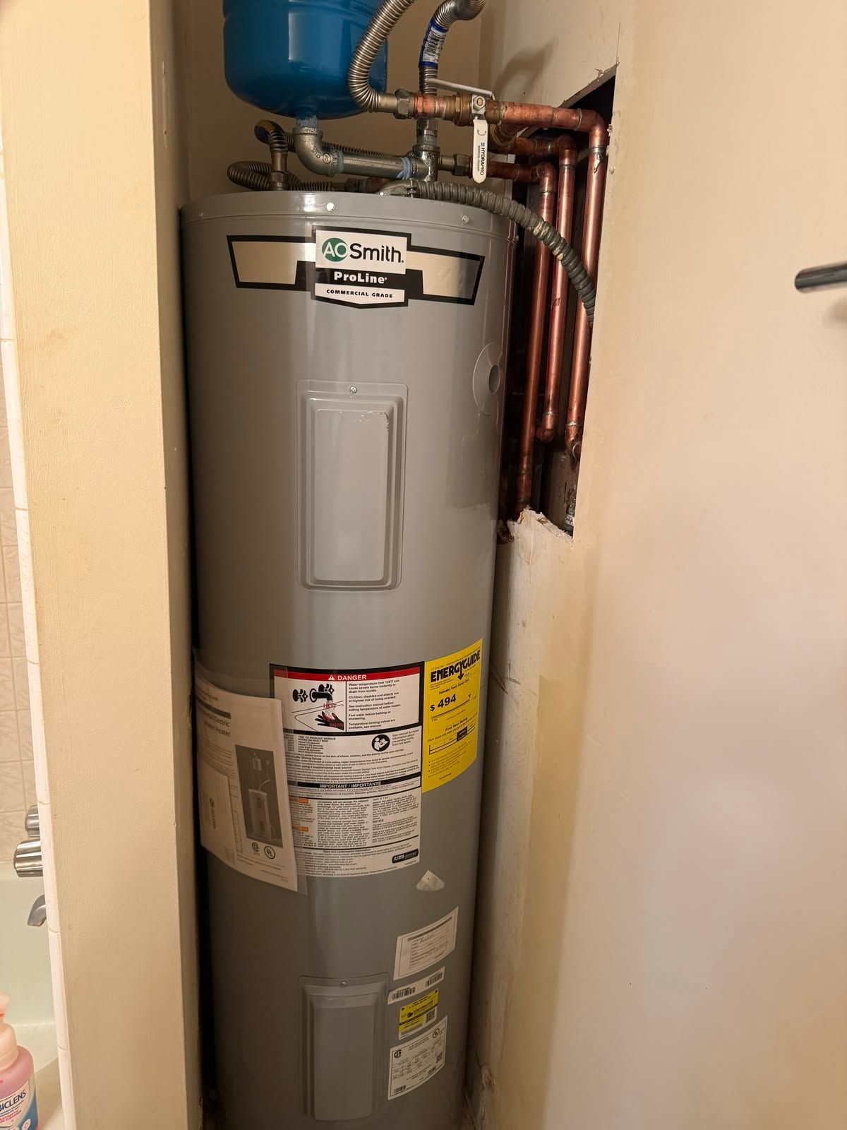Gray water heater in a narrow closet with exposed copper pipes and a blue expansion tank.